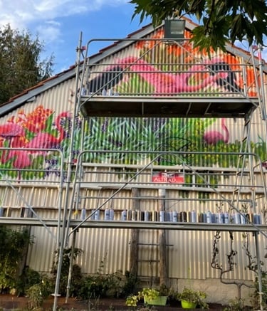 une grange en taule avec un échafaudage et une fresque avec des flamands roses et des plantes
