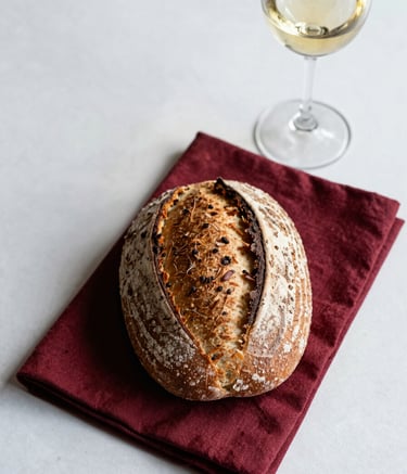 An overhead shot of a beautifully set table in a European / French bistro. A rustic sourdough loaf sits next to a deep red cloth napkin and a glass of wine. The aesthetic is clean and Scandinavian, emphasizing texture and natural ingredients.