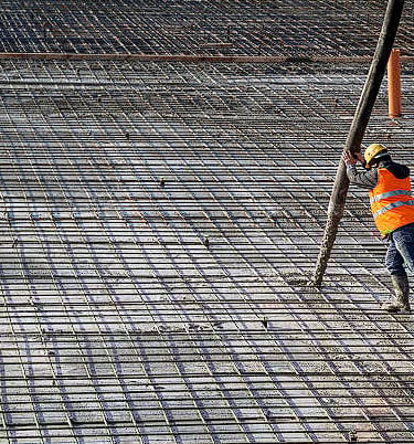 Worker with orange vest and yellow helmet holding boom pump hose for concrete covering wire mesh.