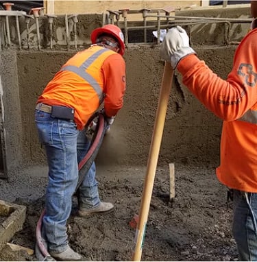 Concrete workers wearing orange safety vests and hard hats, one holds a hose, the other a shovel.