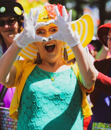 a woman in a green dress with a yellow hat makes a finger heart in front of her face