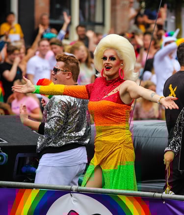 A drag queen in a rainbow dress with large blonde wig holds her arms out in greeting