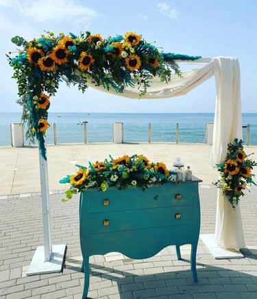 Seaside wedding ceremony setup featuring white arch and teal vintage dresser, adorned with sunflower and turquoise foliage