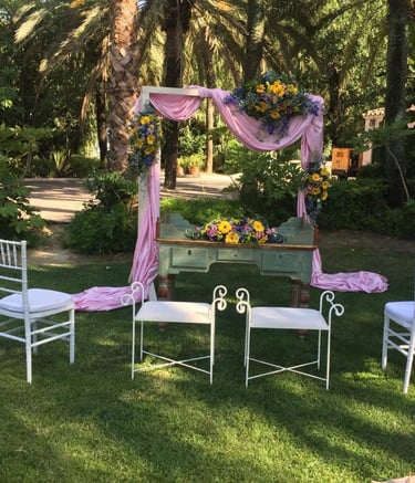 Outdoor wedding ceremony setup in lush garden, pink fabric-draped arch floral arrangements, vintage green desk, white chairs