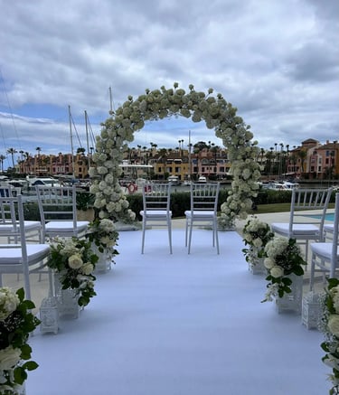 Elegant outdoor wedding ceremony setup featuring a large circular white floral arch, white chairs overlooking a marina.