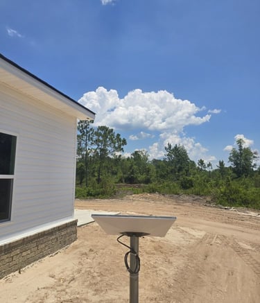 A rectangular Starlink satellite dish mounted on a pole near a rural white house under a bright blue sky.