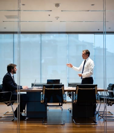 A businessman standing and presenting to a colleague in a modern glass-walled conference room.