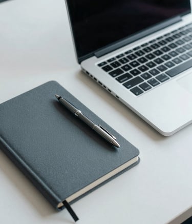 A close-up of a minimalist, organized desk in a North American / International office setting. A modern silver laptop, a charcoal grey notebook, and a premium pen are arranged neatly. The lighting is soft and professional, emphasizing a palette of muted slate blue and soft off-white.