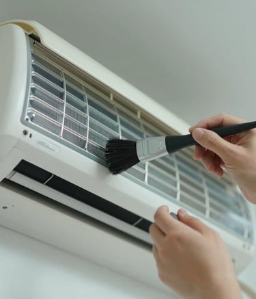 Close-up photography of professional hands using a specialized brush to clean the delicate aluminum fins of an indoor split AC unit, soft light blue and off white tones in the background.