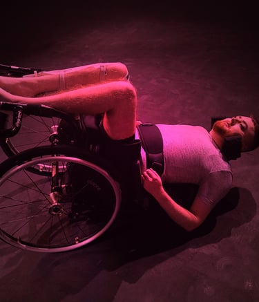 A half-dressed white person in a manual wheelchair on their back on the floor, bathed in pink light.