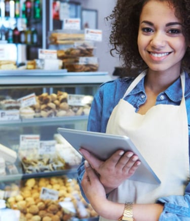 Pessoa em pé  com uma tablet na mão e no fundo uma vitrine de padaria