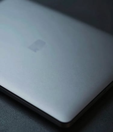 A close-up shot of a sleek, dark metallic laptop lid in a professional Central European / German office setting. The lighting is moody and focused, highlighting the clean lines and texture of the hardware. Colors include Dark Slate Grey and Muted Teal.