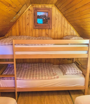 Rustic wooden attic bedroom featuring a light wood bunk bed with plaid bedding and a small window.
