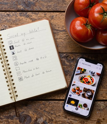 A top-down flat lay on a rustic wooden table in a North American / Western European studio. The scene features a vintage notebook with handwritten social media plans, a bowl of deep ripe crimson tomatoes, and a modern smartphone showing a professional food feed.