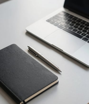 A high-angle, minimalist close-up of a modern professional workspace. A sleek laptop sits on a clean, light grey desk next to a matte black notebook and a silver pen. The scene is bathed in bright, natural morning light, featuring ghost white and muted silver tones.