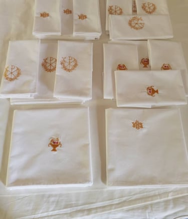 Stacks of white linen altar cloths featuring gold embroidered religious symbols like the IHS and Chi Rho.