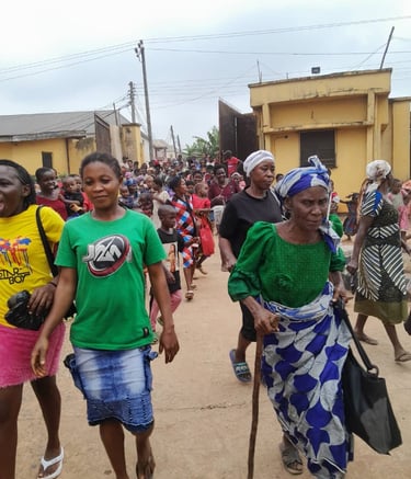 A diverse crowd of people, including an elderly woman with a walking stick, walking down a village street.