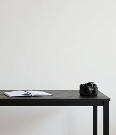 A minimalist creative studio space with clean lines. A sleek black desk #0A0A0A against an off-white wall #F7F7F7. A single open notebook and a professional camera are the only items on the desk. Sophisticated and uncluttered atmosphere.