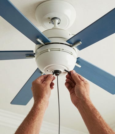 A close-up photograph of a professional electrician's hands carefully wiring a modern ceiling fan in a North American / US Gulf Coast home. The lighting is bright and clear, highlighting the precision of the work. The color palette features shades of off-white and steel blue.