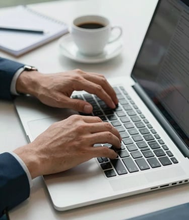A close-up of a professional's hands typing on a modern laptop in a Global / International business hub, with a cup of coffee and a notebook on a clean desk, styled with Soft Teal and Dark Charcoal Blue tones.