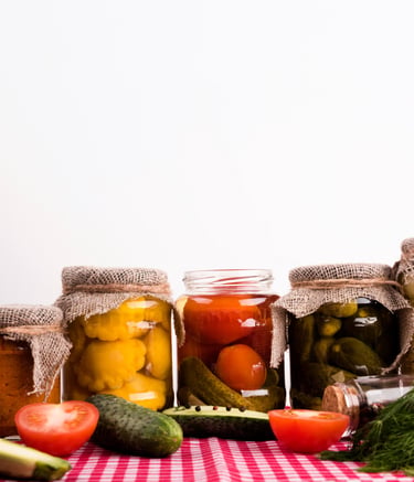 Assorted glass jars of pickled vegetables including cucumbers, tomatoes, and squash on a red checkered cloth.