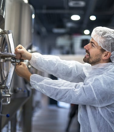 A brewery worker in protective gear adjusts a stainless steel fermentation tank.