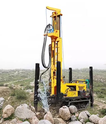 Yellow crawler-mounted hydraulic water well drilling rig operating on a rocky construction site.