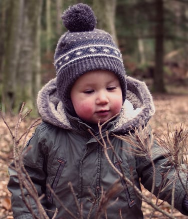 Toddler in a winter hat and warm coat exploring dry branches in a scenic autumn forest.