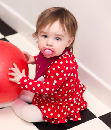 A toddler in a red polka dot dress holding a large red ball on a black and white tiled floor.