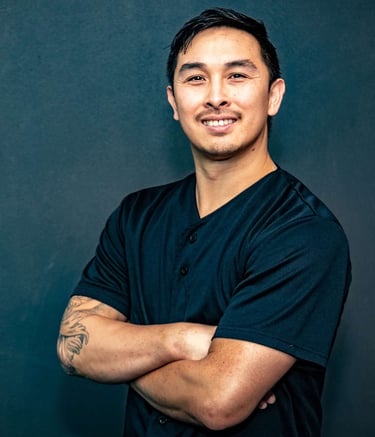 Smiling man with arm tattoos posing in a black shirt against a dark studio background.