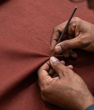 A detailed close-up of a skilled South Asian artisan's hands meticulously stitching heavy-duty upholstery fabric in a muted red-brown color. The lighting is focused and professional, highlighting the texture of the material and the precision of the work.