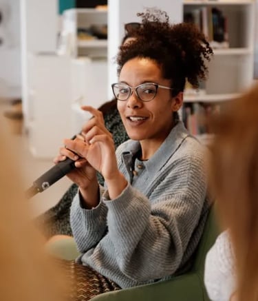 Una mujer con gafas y un suéter sentada hablando a un microfono