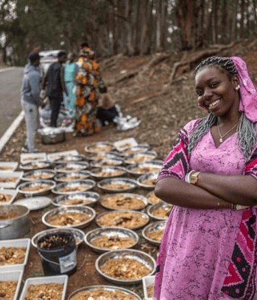Una mujer afro con un vestido rosa parada frente a varios platos llenos de comida.