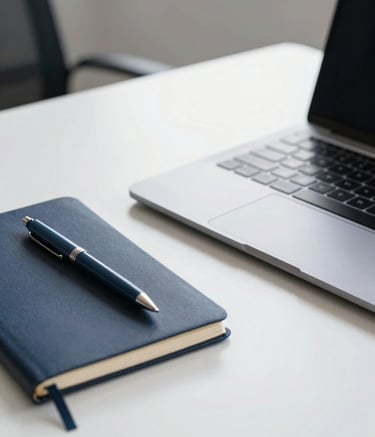 A close-up photograph of a professional's desk in a minimalist office. On the surface sits a sleek metallic laptop and a midnight navy notebook with a slate blue pen. The lighting is bright and natural, reflecting an atmosphere of focused, professional work.