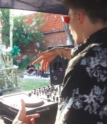 A DJ wearing a floral shirt plays a live set at an outdoor garden party with floating bubbles.