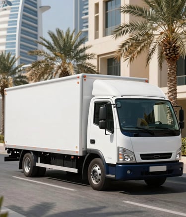 A sleek white and navy blue moving truck parked in a luxury Dubai street with contemporary Middle Eastern / Gulf architecture and palm trees. The bright sun casts crisp shadows on the clean pavement.