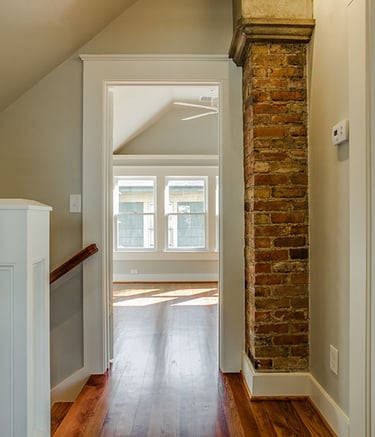 Rustic modern home hallway with exposed brick pillar, hardwood floors, and view into a bright bedroom.