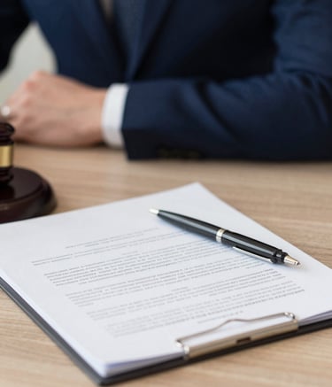 A close-up photograph of professional legal documents and a pen on a clean wooden desk. In the background, out of focus, a person in a deep navy blue blazer is working. The atmosphere is professional, organized, and expert.