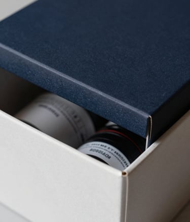 Close-up of a premium, matte-finished delivery box being opened, revealing a glimpse of high-quality goods inside. The scene is shot with a shallow depth of field, using dark navy and off-white tones.