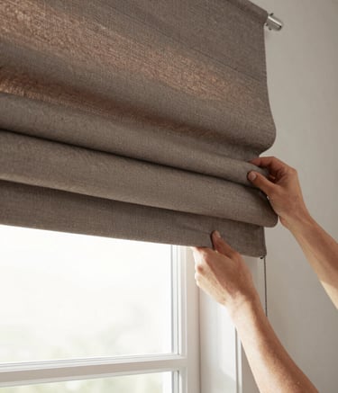 A detailed photography shot of a professional installation of a roman shade in a North American home. The fabric is a muted brown taupe with a clean, modern fold. Soft, diffused lighting highlights the craftsmanship and texture of the textile. Focused composition on the window header and the expert mounting.