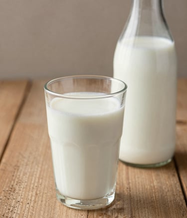 Close-up photography of a fresh glass of milk and a glass bottle on a rustic wooden table, soft morning light, warm beige and muted taupe brown color palette.