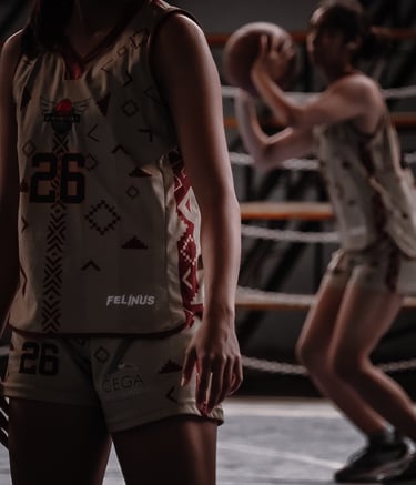 A female basketball player in a custom patterned jersey stands on court while a teammate shoots a ball.