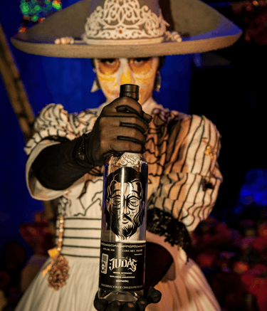 A person in Catrina makeup holding a bottle of Mezcal Judas artisanal Mexican spirit.