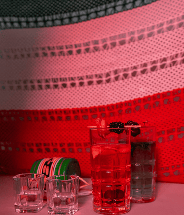 Red craft cocktails and tequila shots garnished with blackberries on a table with a Mexican flag background.