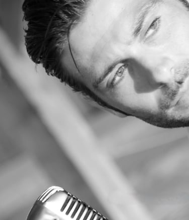 Black and white photo of a handsome man with slicked-back hair looking down at a vintage microphone, perhaps a singer or