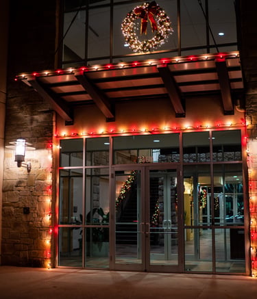 Office building glass entrance decorated with red Christmas lights and a lit holiday wreath at night.