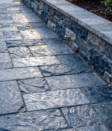A detailed close-up shot of a newly installed flagstone path and a sturdy stone retaining wall featuring dark slate blue tones in a well-maintained North American / US garden. The lighting is soft afternoon sun, emphasizing the professional craftsmanship and textured silver grey surfaces.