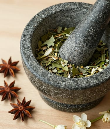 A close-up, overhead shot of a stone mortar and pestle containing crushed green leaves and herbs. Beside it lie whole dried spices and fresh jasmine petals on a light wood surface. The color palette includes Muted Moss Green and Warm Off-White. The lighting is soft morning sun, creating a serene and natural atmosphere.