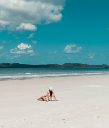 A woman wearing a sun hat sits on a white sand tropical beach with blue ocean water and a cloudy sky.