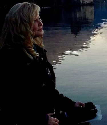 A blonde woman looking at a peaceful sunset reflection on a lake near a historic castle.